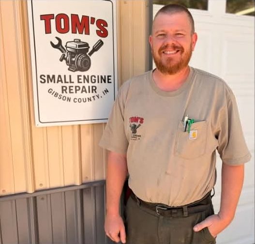 Tom Atkinson standing beside the Tom's Small Engine Repair sign on his shop.